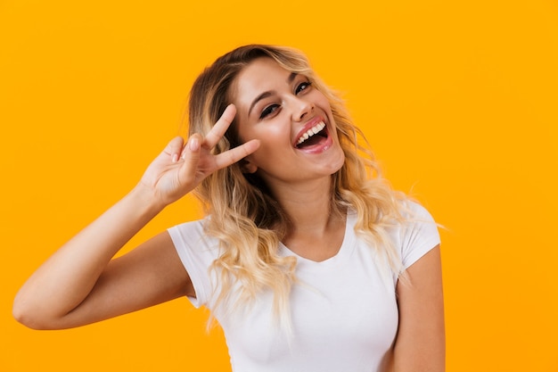 Blond woman in basic clothing smiling and showing peace sign, isolated over yellow wall