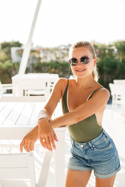 Blond woman beautiful face tanned skin dressed in green top and denim shorts fashion clothing style summer clothes collection summer and leisure