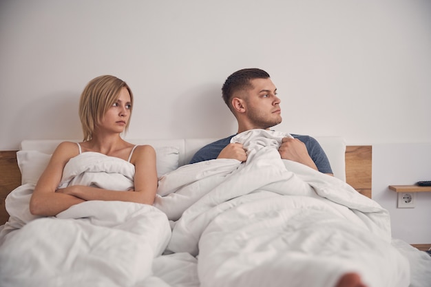  blond woman and brunette male staying in bed with white linen while both looking disappointed