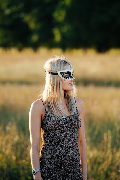 Blond woman in a mask and a glittering dress in front of a dry grass field