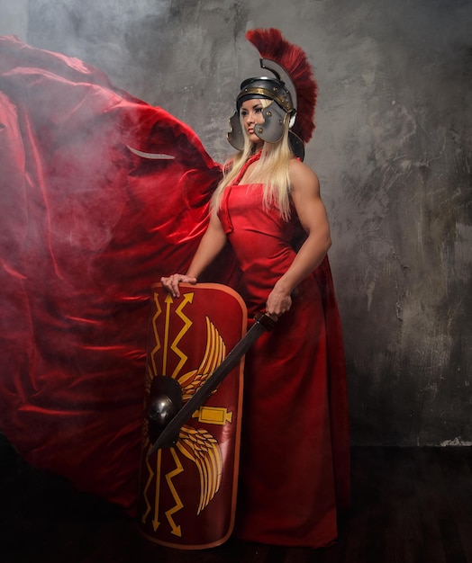 Blond woman in Roman red fluttering dress holds sword and shield.