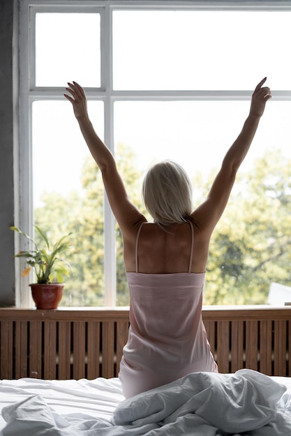 Blond woman stretches in the morning on the bed raising her hands up Slim millennial in silk lingerie in the bedroom start of day weekend Beauty and health concept View back