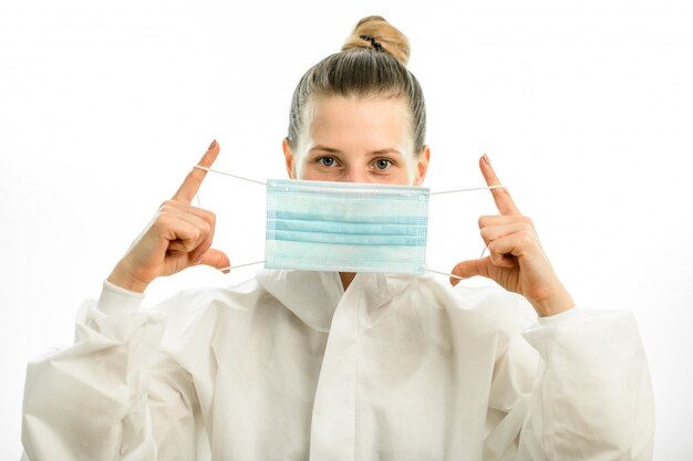 Blond woman in white protective suit holds medical mask in front of her face