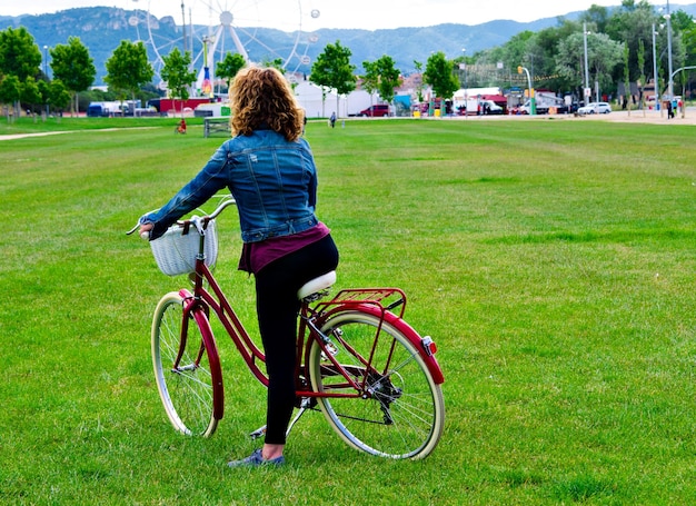 Blond woman with blue denim strolling in the park on a red bicycle.