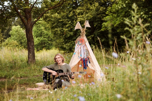 Blonde authentic woman is playing the guitar next to wigwam decorated with dream catchers outdoor.