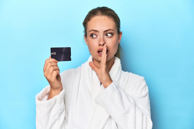 Blonde in bathrobe with credit card in blue studio is saying a secret hot breaking news