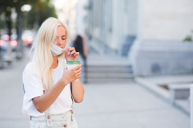 Blonde beautiful womanholding paper coffee cup and wearing medical mask