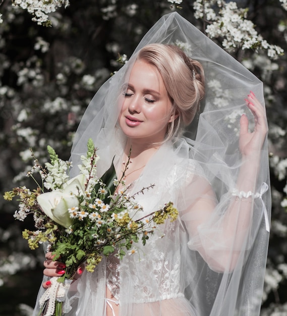 A blonde bride with a veil and a bouquet of flowers poses in a blooming garden