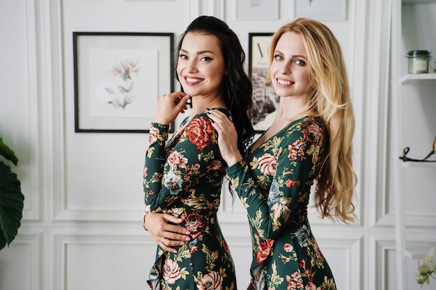 Blonde and brunette in short green dresses with patterns. Two beautiful girls in a white studio.