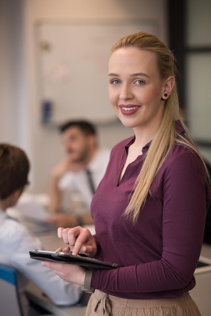 Blonde businesswoman in casual  clothes working on tablet computer at modern startup business office interior. Young people group on team meeting blured  in background