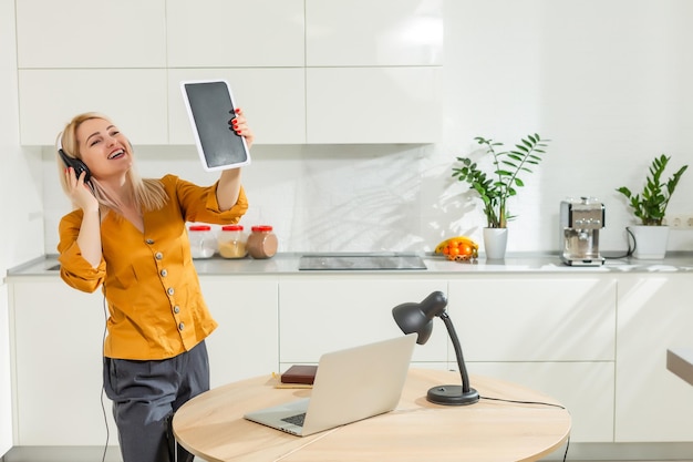 blonde caucasian girl woman working from home at a table in the kitchen in using a book a pen a table with candles busy office lock down covid tablet in her hand