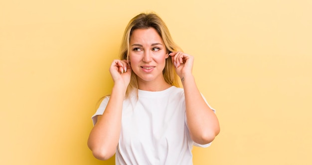 Blonde caucasian woman looking angry stressed and annoyed covering both ears to a deafening noise sound or loud music