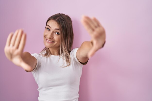 Blonde caucasian woman standing over pink background doing frame using hands palms and fingers, camera perspective