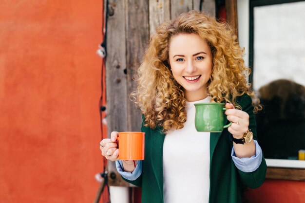 Blonde curlyhaired woman with coffee mugs happy expression pleasing smile ready to treat friends