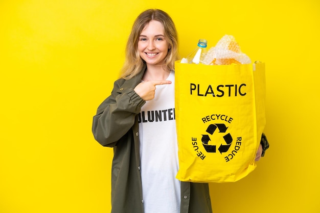 Blonde English young girl holding a bag full of plastic bottles to recycle isolated on yellow background and pointing it