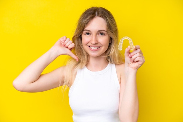 Blonde English young girl holding invisible braces isolated on yellow background proud and selfsatisfied