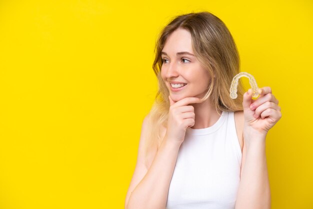 Blonde English young girl holding invisible braces isolated on yellow background thinking an idea and looking side