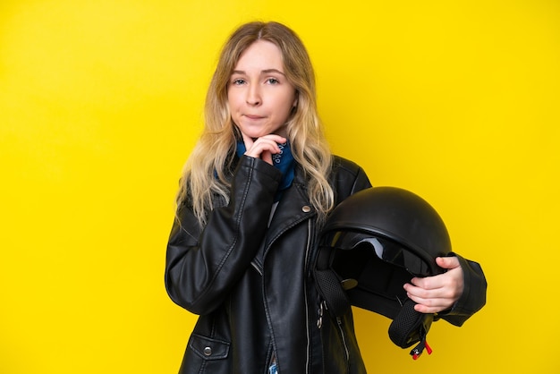 Blonde English young girl with a motorcycle helmet isolated on yellow background having doubts and thinking