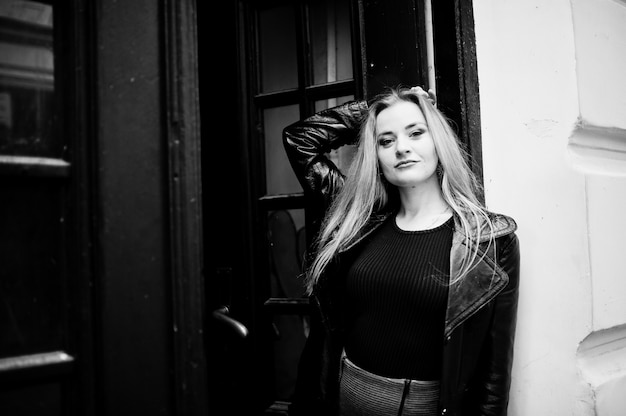 Blonde fashionable girl in long black leather coat posed against wooden door at old house.