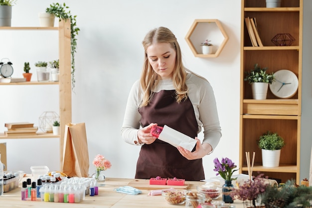 Blonde female in apron putting handmade soap bar into paper packet to give it to someone as gift for holiday