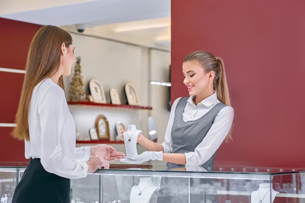 Blonde female worker in a grey uniform showing a necklace to a female client in a jewelry store