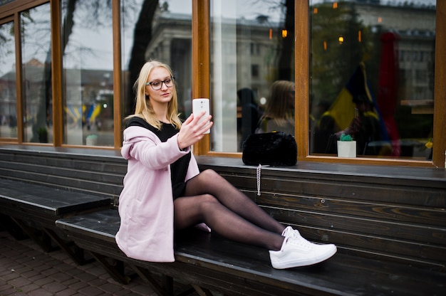 Blonde girl at glasses and pink coat, black tunic sitting on bench at street and doing selfie from smartphone.