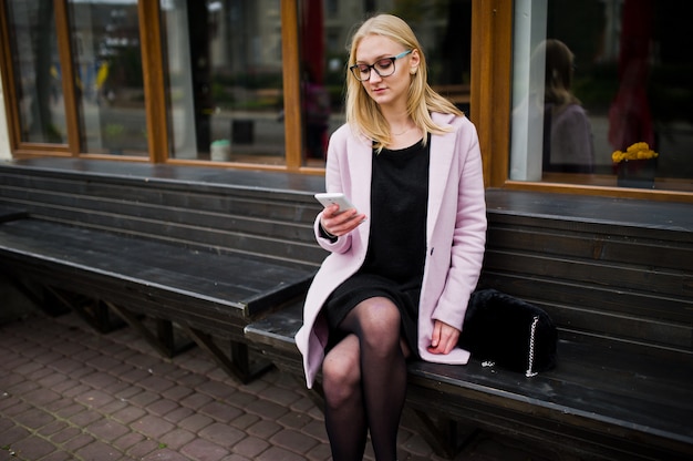 Blonde girl at glasses and pink coat, black tunic sitting on bench at street and looking on smartphone.