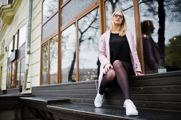 Blonde girl at glasses and pink coat, black tunic sitting on bench at street.