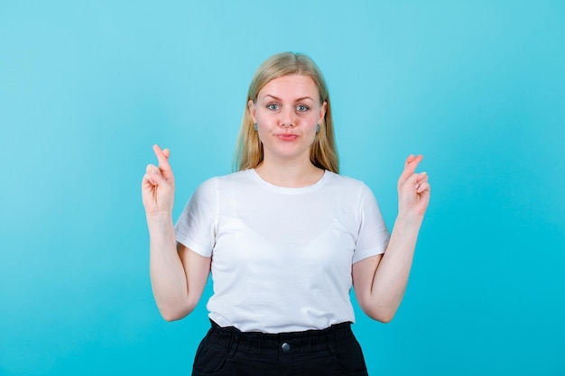 Blonde girl is looking at camera by crossing and raising up her fingers on blue background