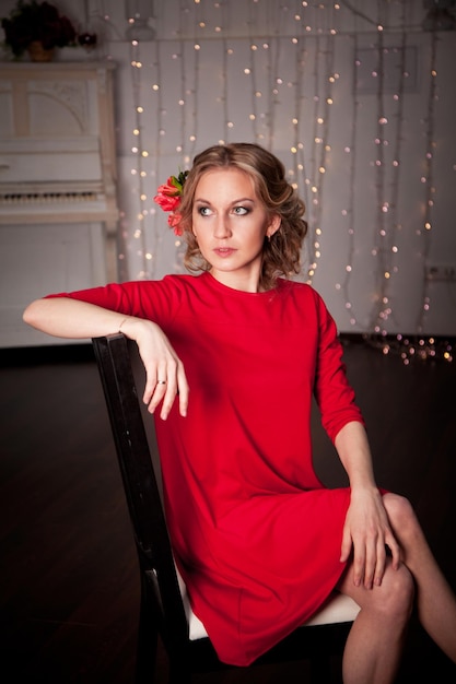 A blonde girl in a red dress and with flowers in her hair sits on a leather sofa