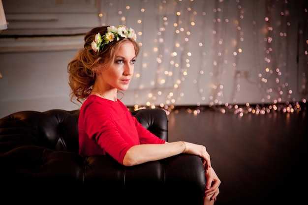 A blonde girl in a red dress and with flowers in her hair sits on a leather sofa