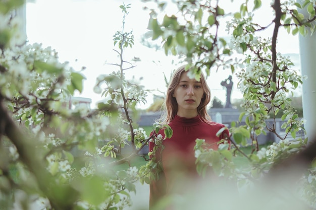 Photo a blonde girl in red looks into the camera standing behind a flowering tree spring sunny