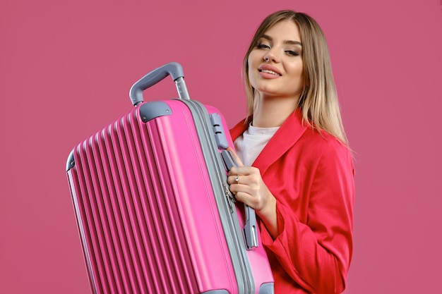 Blonde girl in red pantsuit and white blouse she smiling holding suitcase posing on pink background