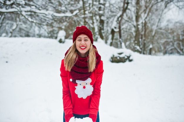 Blonde girl in red scarf, hat and santas sweater posing at park on winter day.