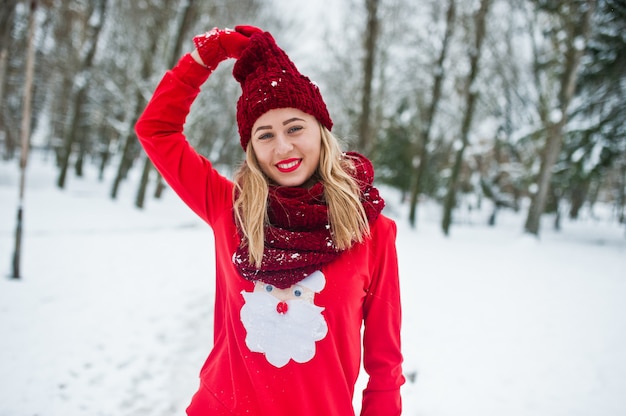 Blonde girl in red scarf, hat and santas sweater posing at park on winter day.