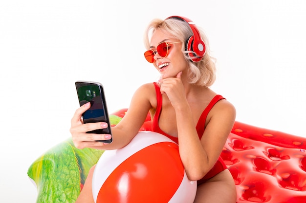 blonde girl in a red swimsuit and sunglasses sitting on swimming mattress and listening to music on headphones and holding a smartphone in her hands