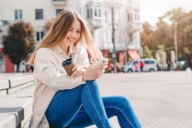 Blonde girl sitting on the steps with a mobile phone laughing and texting with the messenger