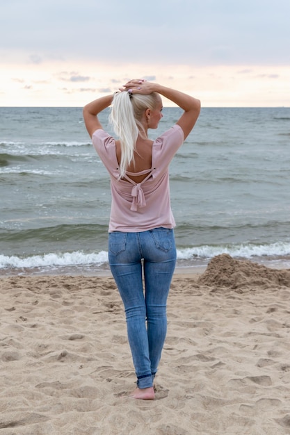 A blonde girl stands on the beach by the sea with her hands behind her head and looks at the sunset