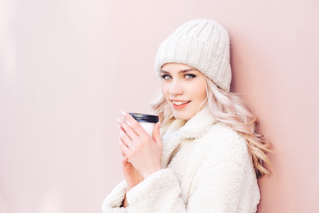 The blonde girl in winter clothes is holding a coffee in a paper cup on a pink background. Young woman smiling and looking at the camera.