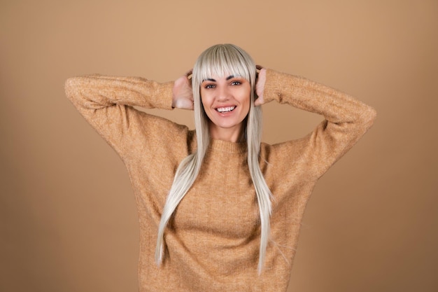 A blonde girl with bangs and brown daytime makeup in a sweater on a beige background laughs merrily, covering her ears with her hands