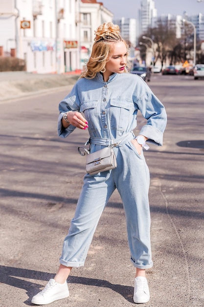 Photo blonde girl with bright makeup and hairstyle in denim jumpsuit posing on a city street