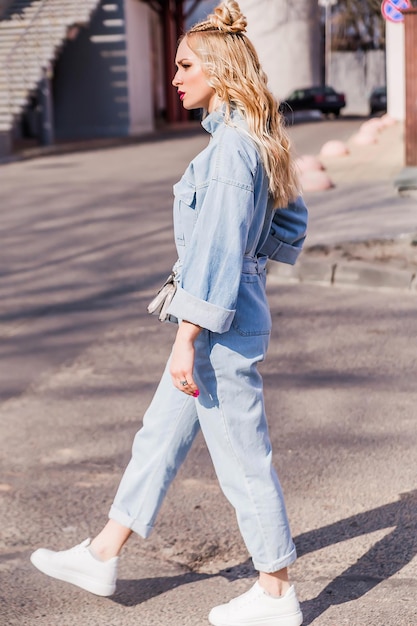 Blonde girl with bright makeup and hairstyle in denim jumpsuit posing on a city street