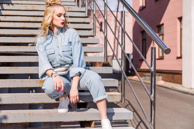 A blonde girl with bright makeup and a hairstyle in denim overalls sits on the stairs on the street