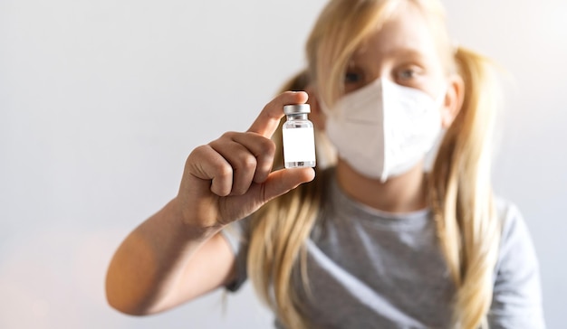 blonde girl with face mask shows a Vaccine bottle for Covid-19 vaccine with white label with copy space for text at a vaccination center.