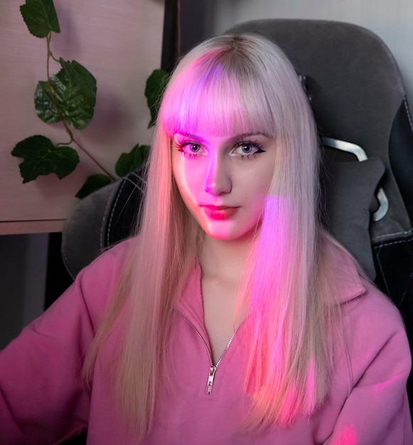 A blonde girl with long hair and straight bangs in a pink blouse is smiling in her room in a computer chair