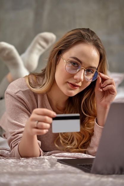 Blonde lady blogger in glasses casual clothing showing plastic card lying crosslegged on bed with la...