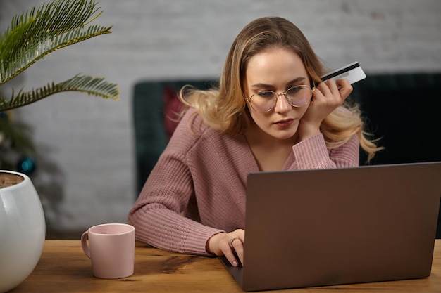 Blonde lady in pink cardigan and glasses. Working on her laptop and holding white plastic card. Sitting at wooden table with pink cup, palm in pot on it. Student, blogger. Home interior. Close up