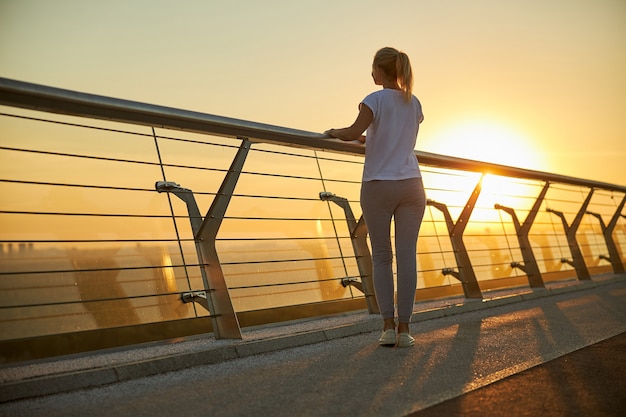 Blonde lady in sportswear enjoying the view of bright sunrise while spending time outdoors