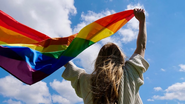 Blonde lesbian woman holding a rainbow lgbt gender identity flag on sky background with clouds on a