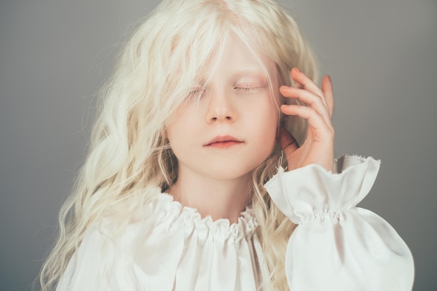 Blonde little girl. Angel beauty. Child purity. Portrait of adorable cute peaceful kid with curly hair closed eyes in white vintage blouse isolated on gray background.